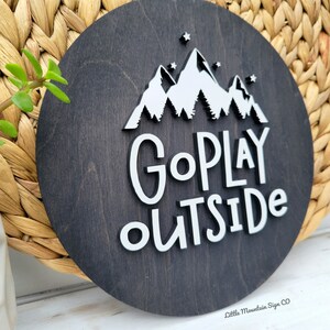May include: A round wooden sign with a dark brown stain. The sign features white 3D lettering that reads "Go Play Outside" with a mountain range and stars behind the text.