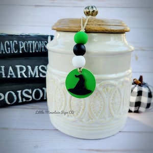 May include: White ceramic jar with a wooden lid and a green, black, and white beaded charm with a witch hat silhouette. The jar has a decorative design and the charm says "Little Mountain Sign Co."