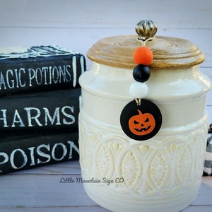 May include: White ceramic jar with a wooden lid and a black and orange Halloween pumpkin charm. The jar has a decorative design and is sitting on a white wooden surface. The charm has a black pumpkin with an orange face and a black stem.