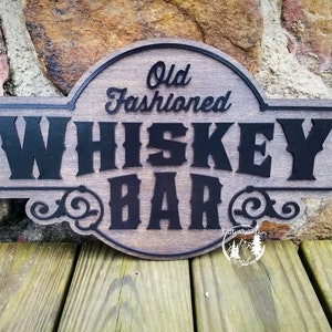 May include: Wooden sign with the words "Old Fashioned Whiskey Bar" in black, stylized lettering. The sign has a dark wood finish and decorative flourishes. The sign is displayed against a stone wall and wooden planks.