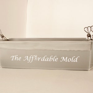 Può includere: Uno stampo in plastica grigia per realizzare vasi di cemento, tenuto insieme da clip metalliche. Lo stampo è etichettato "The Affordable Mold".