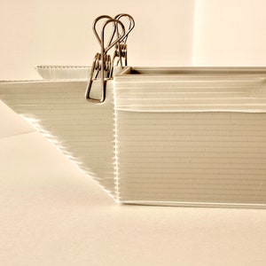 May include: A white cardboard box with a corrugated texture, held together by silver binder clips. The box is open at the top and has a slight curve at the front.