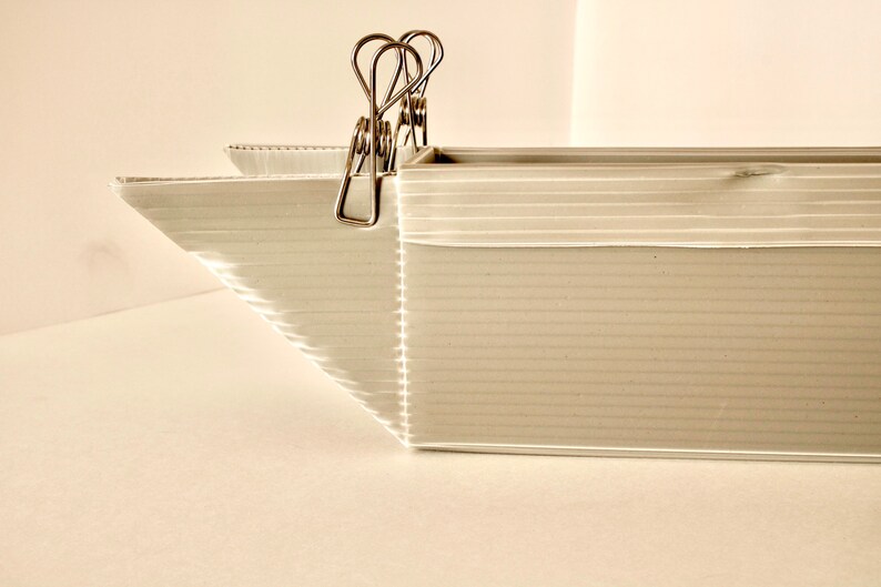 May include: A white cardboard box with a corrugated texture, held together by silver binder clips. The box is open at the top and has a slight curve at the front.