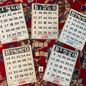 Pu&ograve; includere: Cinque carte da bingo vintage con testo in bianco e nero e numeri rossi. Le carte sono su un tappeto a motivi rossi e blu. Le carte sono della Chiesa di San Giovanni e hanno numeri diversi su ogni carta. Le carte sono per un gioco di bingo.