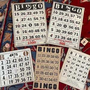 Pu&ograve; includere: Sei cartelle del bingo con numeri da 1 a 75. Le cartelle sono bianche con numeri neri e un titolo nero "Bingo". Le cartelle sono su una superficie con un motivo rosso e dorato.
