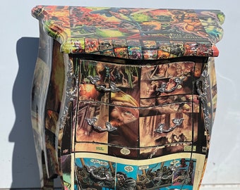 May include: A small, three-drawer nightstand with a unique design. The nightstand is covered in a collage of comic book pages, creating a colorful and eclectic look. The drawers have silver handles and the legs are curved and ornate.