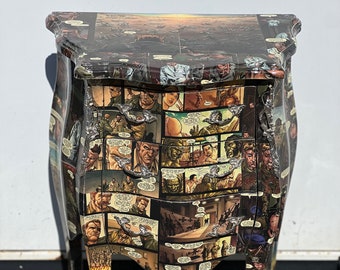May include: A small, three-drawer side table with a unique design. The table is covered in a collage of comic book pages featuring characters and scenes from a war story. The table has ornate, curved legs and silver-toned drawer pulls.