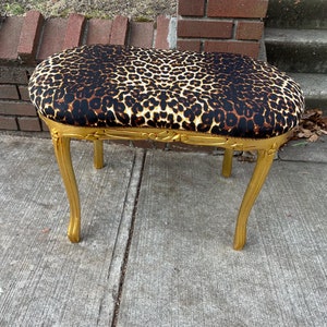 May include: A gold-painted wooden bench with a leopard print upholstered seat. The bench has a curved, oval-shaped seat and ornate legs.