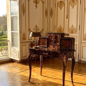 May include: A vintage desk with a leopard print finish. The desk has curved legs and a matching hutch with multiple compartments. A brass lamp sits on the desk, and the room features ornate gold trim and a parquet floor.