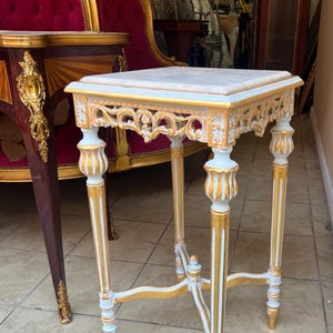 May include: A small, ornate side table with a square marble top. The table features gold and light blue painted details, including decorative carvings along the edges and on the legs. The table is approximately 24 inches tall.