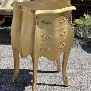 May include: A gold-painted wooden chest of drawers with three drawers and ornate brass handles. The chest is on a light gray surface.