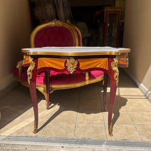 May include: An ornate, antique table with a white marble top and a rich, dark wood finish. The table features gold accents, including decorative carvings on the legs and apron. The legs are curved, and the table has a classic, elegant design.