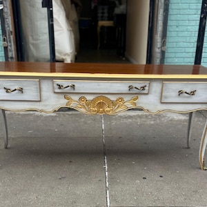 May include: A white and gold console table with three drawers and ornate carvings. The table has a distressed white finish and gold accents. The table is made of wood and has a rectangular shape.