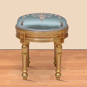 May include: A round, light blue, embroidered ottoman with gold trim and three gold legs. The ottoman is sitting on a wooden floor.