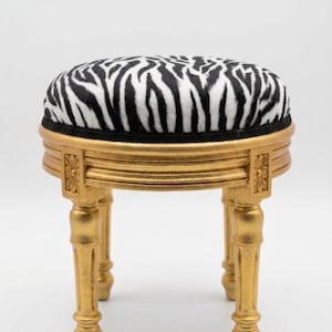 May include: A round stool featuring a black and white zebra-patterned upholstered seat cushion. The base is constructed from gold-painted wood, showcasing four carved legs with decorative square accents at the top of each leg support.