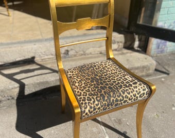 May include: A gold-painted wooden chair featuring a square seat upholstered in a brown and black leopard print fabric. The chair has a curved top rail, a horizontal back splat, and four legs, with decorative nailhead trim along the seat edge.