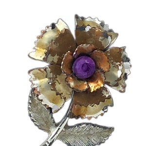 Op de afbeelding: Een vintage bloem broche met een paarse strass steentje in het midden. De bloem is gemaakt van metaal met een gouden en zilveren afwerking.