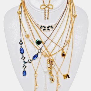 May include: A display of necklaces on a white bust. The necklaces include gold chains, blue and clear gemstones, pearls, and a gold initial "M". Some necklaces have bow and teardrop pendants. Various lengths and styles are shown.