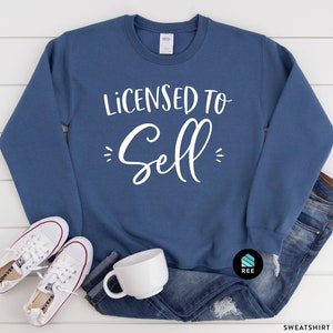 May include: A blue sweatshirt with the text "Licensed to Sell" in white lettering. The sweatshirt is laying on a white wooden surface with a pair of white sneakers and a white mug.