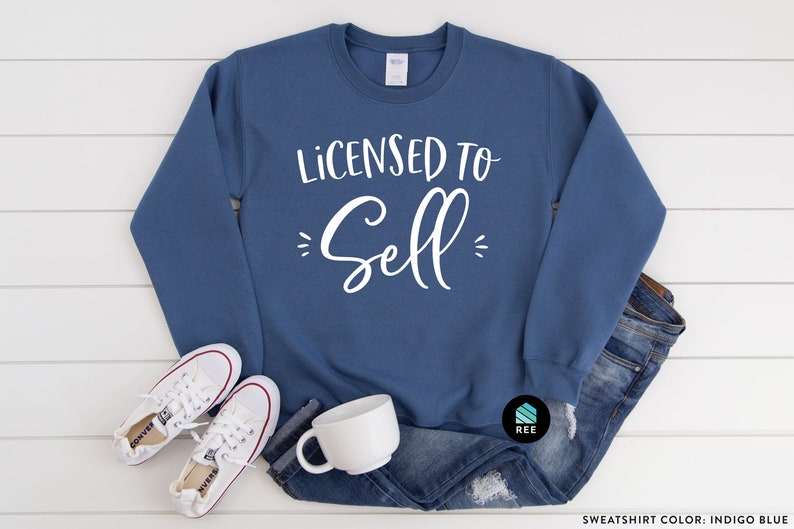 May include: A blue sweatshirt with the text "Licensed to Sell" in white lettering. The sweatshirt is laying on a white wooden surface with a pair of white sneakers and a white mug.