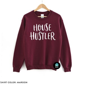Peut inclure: Sweat-shirt bordeaux avec l'inscription blanche &laquo; HOUSE HUSTLER &raquo;. Le sweat-shirt est suspendu &agrave; un cintre en bois sur fond blanc. Col rond, couleur unie.