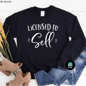 May include: A black sweatshirt with the text "Licensed to Sell" in white lettering. The sweatshirt is laid out on a white wooden surface with a pair of brown heeled shoes and blue jeans.