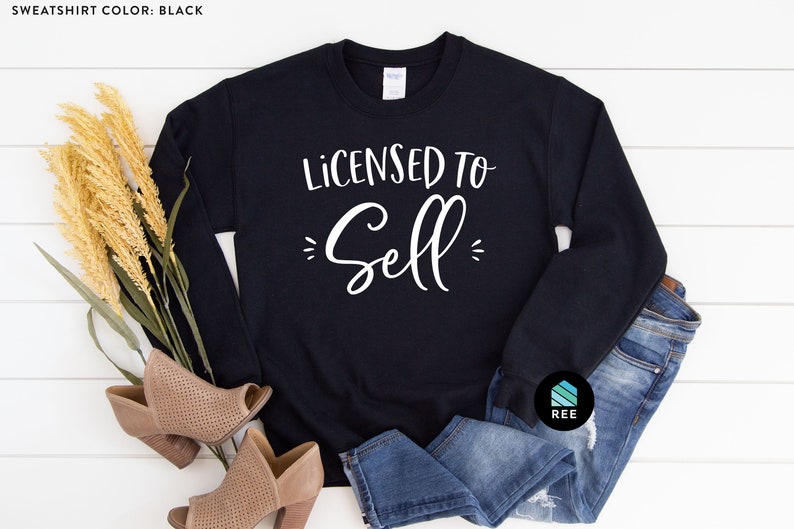May include: A black sweatshirt with the text "Licensed to Sell" in white lettering. The sweatshirt is laid out on a white wooden surface with a pair of brown heeled shoes and blue jeans.