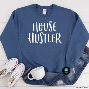 Peut inclure: Sweat-shirt bleu indigo avec le texte "HOUSE HUSTLER" en blanc. Le sweat-shirt est associ&eacute; &agrave; des baskets blanches, une tasse blanche et un jean bleu. Le sweat-shirt est de style col rond.
