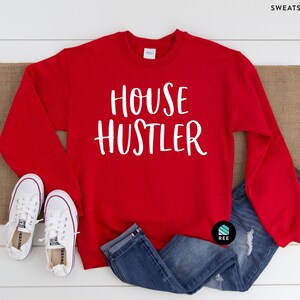 Peut inclure: Un sweat-shirt rouge avec le texte blanc "HOUSE HUSTLER" est pr&eacute;sent&eacute; sur une surface en bois blanc. Le sweat-shirt est associ&eacute; &agrave; un jean bleu et &agrave; des baskets blanches. Un chemin de table en toile de jute marron est sous le sweat-shirt.
