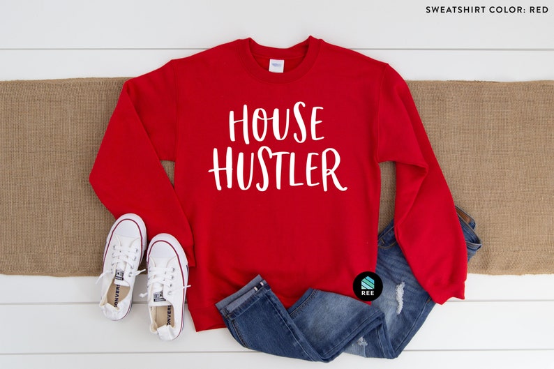 Peut inclure: Un sweat-shirt rouge avec le texte blanc "HOUSE HUSTLER" est pr&eacute;sent&eacute; sur une surface en bois blanc. Le sweat-shirt est associ&eacute; &agrave; un jean bleu et &agrave; des baskets blanches. Un chemin de table en toile de jute marron est sous le sweat-shirt.