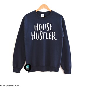 Peut inclure: Sweat-shirt bleu marine avec le texte blanc "HOUSE HUSTLER". Le sweat-shirt est suspendu &agrave; un cintre en bois. Les mots "SWEATSHIRT COLOR: NAVY" sont au bas de l'image.