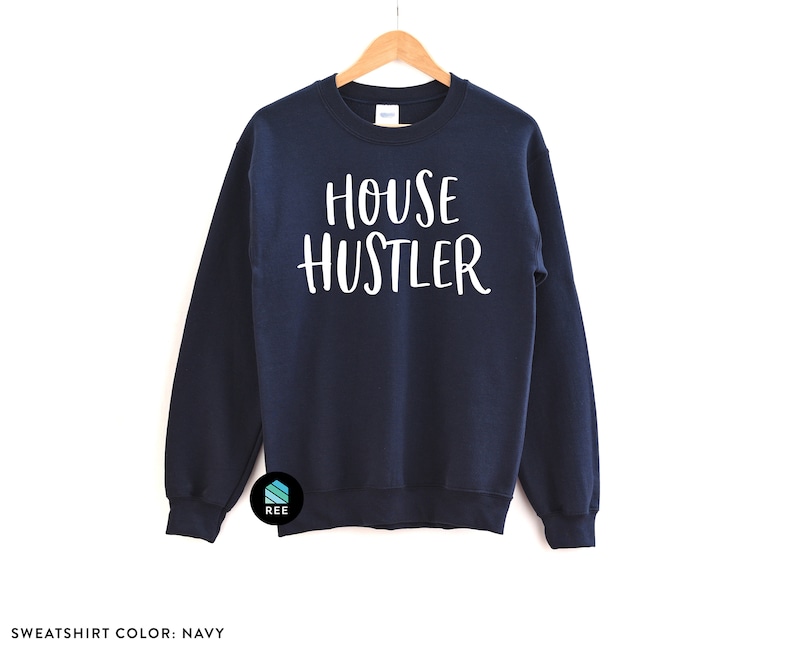 Peut inclure: Sweat-shirt bleu marine avec le texte blanc "HOUSE HUSTLER". Le sweat-shirt est suspendu &agrave; un cintre en bois. Les mots "SWEATSHIRT COLOR: NAVY" sont au bas de l'image.