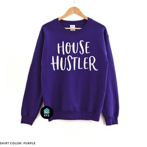 Peut inclure: Un sweat-shirt violet avec l'inscription "HOUSE HUSTLER" en blanc. Le sweat-shirt est suspendu &agrave; un cintre en bois sur fond blanc. Le texte "SWEATSHIRT COLOR: PURPLE" est en bas.