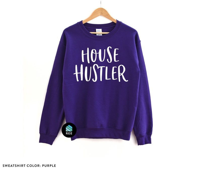 Peut inclure: Un sweat-shirt violet avec l'inscription "HOUSE HUSTLER" en blanc. Le sweat-shirt est suspendu &agrave; un cintre en bois sur fond blanc. Le texte "SWEATSHIRT COLOR: PURPLE" est en bas.