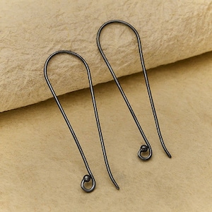Könnte beinhalten: Ein Paar dunkelgraue Metallohrringe. Die Ohrringe haben ein einfaches, längliches Haken-Design mit einem kleinen, abgerundeten Detail am unteren Ende. Sie sind vor einem neutralen, strukturierten Hintergrund abgebildet.