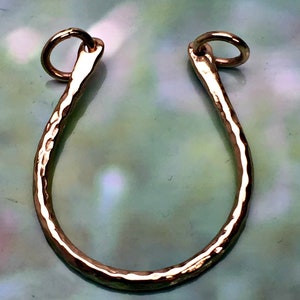 May include: A gold-colored, hammered metal pendant in a U-shape with a small ring at each end. The pendant has a textured surface and is likely for jewelry making or as a decorative element.