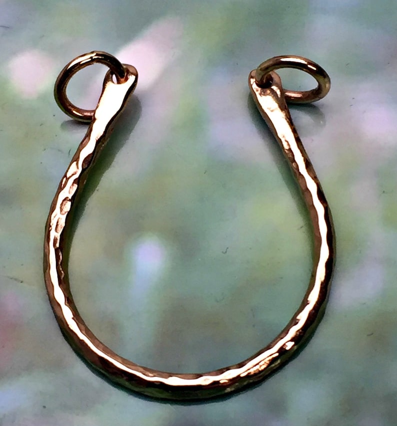 May include: A gold-colored, hammered metal pendant in a U-shape with a small ring at each end. The pendant has a textured surface and is likely for jewelry making or as a decorative element.