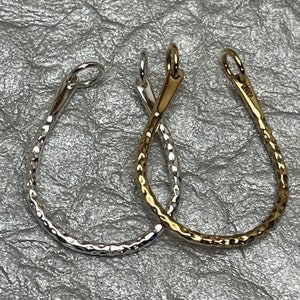 May include: Two teardrop-shaped pendants. One is silver-colored, the other is gold-colored. Both have a hammered texture and a small loop at the top for hanging. The pendants are displayed on a textured, silver background.