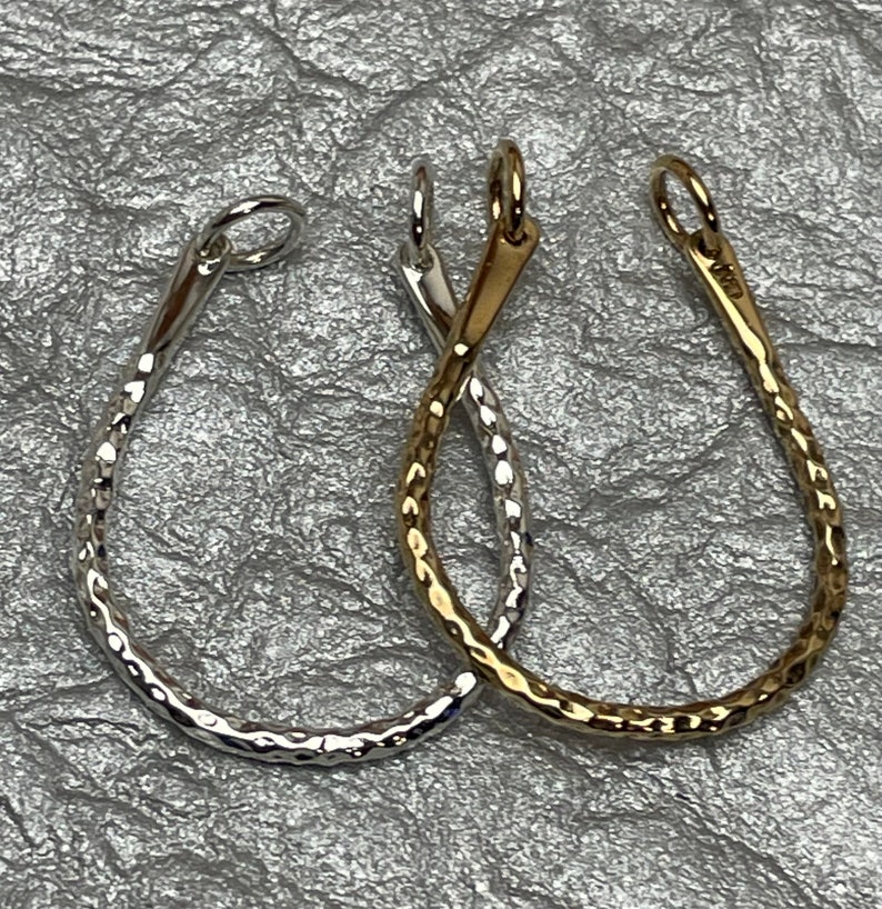 May include: Two teardrop-shaped pendants. One is silver-colored, the other is gold-colored. Both have a hammered texture and a small loop at the top for hanging. The pendants are displayed on a textured, silver background.