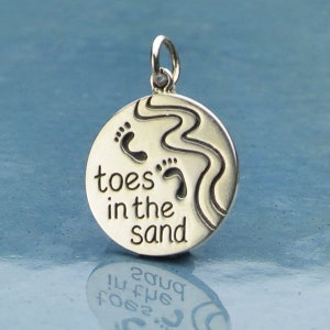 May include: Silver charm with the words "toes in the sand" and two footprints, a wavy line representing water, and a loop for attaching to a necklace or bracelet.