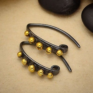 May include: A pair of handcrafted earrings with a dark grey, curved design. Each earring features multiple small, gold-coloured spheres along the curve. The earrings are displayed on a neutral-toned surface.