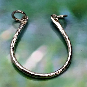 May include: A silver-colored, hammered metal horseshoe-shaped charm with a small loop at each end. The charm has a textured surface and is designed to be worn as a pendant or used in jewelry making.