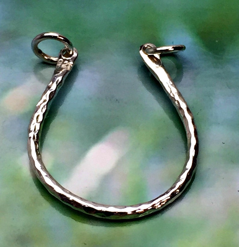 May include: A silver-colored, hammered metal horseshoe-shaped charm with a small loop at each end. The charm has a textured surface and is designed to be worn as a pendant or used in jewelry making.