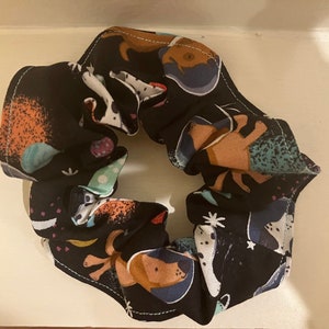 May include: A black scrunchie with a colorful pattern of dogs and abstract shapes. The scrunchie is made of fabric and has a gathered design.