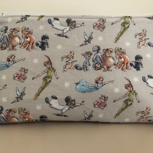 Puede incluir: Un neceser rectangular de tela gris con cremallera blanca. La bolsa está estampada con ilustraciones de Peter Pan, Wendy, Campanilla y otros personajes de la historia clásica, junto con estrellas blancas.