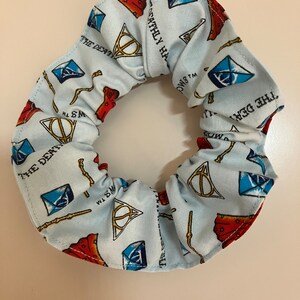 May include: A light blue scrunchie with a repeating pattern of the Deathly Hallows symbol, a wand, and a blue diamond. The text "The Deathly Hallows" is repeated throughout the pattern.