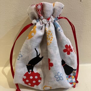 May include: A small, white drawstring bag with a repeating pattern of black cats, red flowers, and yellow and blue shapes. The bag is tied with a red ribbon.
