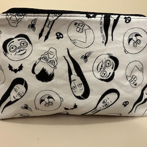 May include: A white zippered pouch with a black and white print of characters from the Addams Family television show. The print includes Gomez, Morticia, Wednesday, Pugsley, and Uncle Fester. The text "©2022 MGM TM TeKa" is printed on the pouch.