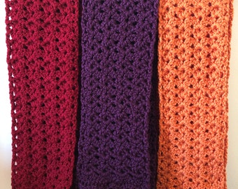 May include: Three crocheted scarves in different colors: burgundy, purple, and orange. The scarves are hanging on a white wall.