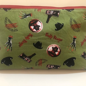 Puede incluir: Bolsa verde con cremallera con un patrón repetido del guante de Freddy Krueger, un sombrero fedora negro y las palabras "Springwood" y "Elm St."